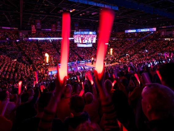 A crowd of people attend of basketball game in an indoor arena. The crowd is holding red, illuminated plastic sticks.