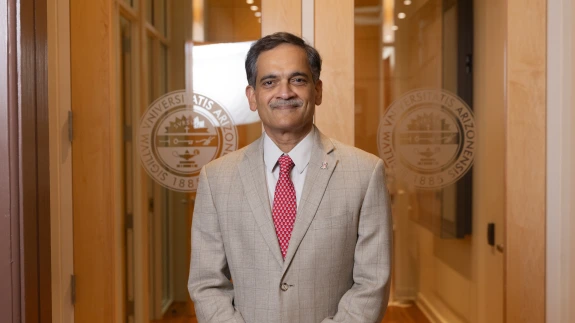 Suresh Garimella standing in front of a glass doorway with the seal of the University of Arizona over each shoulder and a wood-paneled hallway behind him. 
