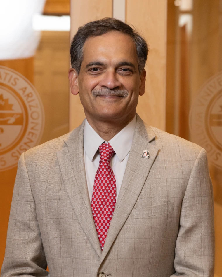 Suresh Garimella standing in front of a glass doorway with the seal of the University of Arizona over each shoulder and a wood-paneled hallway behind him. 