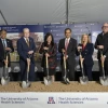 A group of six people stands under a tent, each holding a ceremonial shovel in preparation for a groundbreaking event. They are standing in front of a banner with the University of Arizona Health Sciences logo and a rendering of a new facility. Each person is dressed in business attire, and they smile as they pose for the photo. Helmets are lined up on the stage behind them.