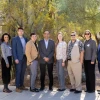 This image shows a group of nine individuals standing outdoors, posing for a photo in front of lush desert vegetation including cacti and other greenery. They are dressed in professional and business-casual attire, smiling and looking at the camera in bright daylight.