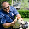 A man wearing sunglasses and a navy blue polo shirtkneels next to a medium-sized yellow dog wearing a "Service Dog" vest.